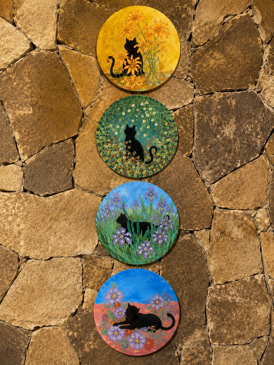 Four round cat paintings displayed vertically on a stone wall from the Flavia and Matteo collaboration
